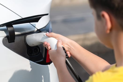 Kid's hand insert EV charger plug into electric vehicle, recharge EV car battery from outdoor charging station. Fresh daylight with alternative clean and sustainable energy concept. Perpetual