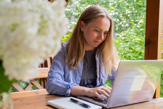 Young happy woman focuses on her laptop in wooden alcove. Relaxed outdoor setting emphasizes comfort and productivity. Remote work learning concept