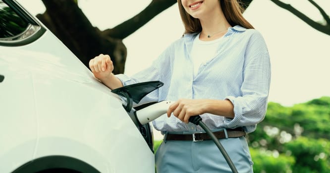 Young woman recharging battery for electric car during road trip travel EV car in natural forest or national park. Eco friendly travel during vacation and holiday. Exalt