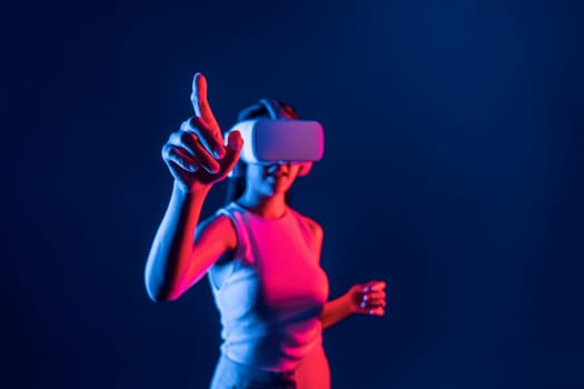 Smart female standing in cyberpunk neon light wear VR headset connecting metaverse, futuristic cyberspace community technology. Elegant woman use finger point generated virtual object. Hallucination.