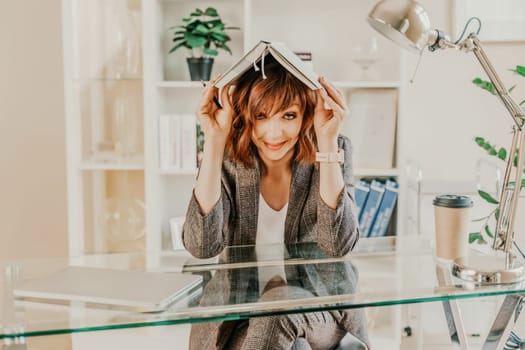 Woman book on her head. Surprised happy brunette in a jacket with a book on her head looks away against the backdrop of a bright office