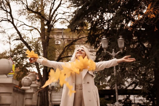 A woman is standing in the street, holding a bunch of yellow leaves and smiling. The leaves are falling from the trees and the woman is enjoying the moment