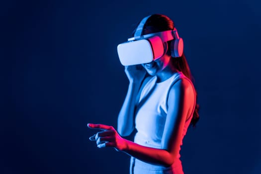 Smart female stand with surrounded by cyberpunk neon light wear VR headset connecting metaverse, futuristic cyberspace community technology. Woman using finger pointing virtual object. Hallucination.