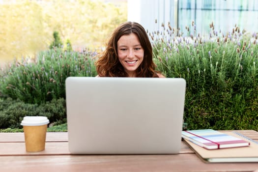 Front view of happy redhead female college student using laptop outdoors sitting on table studying and doing homework. University student and technology concept.