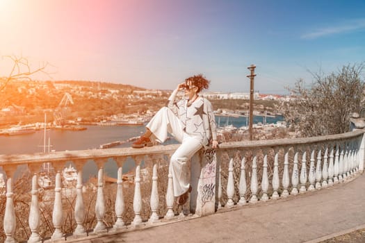 Woman walks around the city, lifestyle. A young beautiful woman in white trousers and a sweater sits on a white fence with balusters and overlooks the sea bay and the city