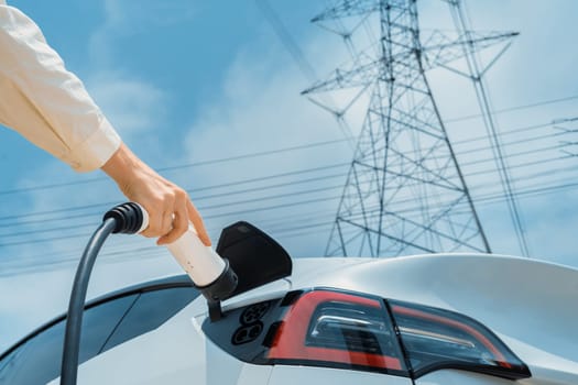 Closeup woman recharge EV electric car battery at charging station connected to electrical power grid tower on sky background as electrical industry for eco friendly vehicle utilization. Expedient