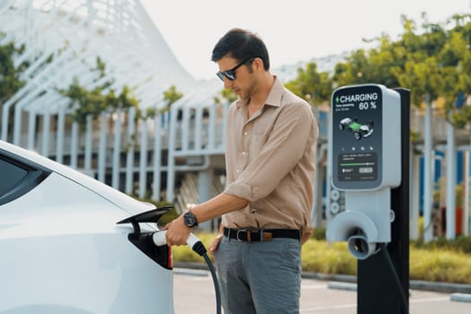 Young man put EV charger to recharge electric car's battery from charging station in city commercial parking lot. Rechargeable EV car for sustainable environmental friendly urban travel. Expedient