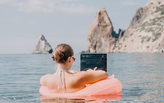 Woman freelancer works on laptop swimming in sea on pink inflatable ring. Pretty lady typing on computer while floating in the sea on inflatable donut at sunset. Freelance, remote work on vacation