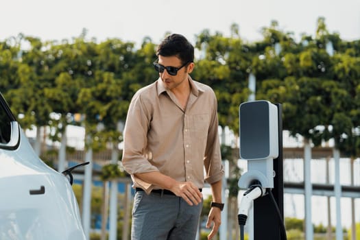 Young man recharge electric car's battery from charging station in outdoor green city park in springtime. Rechargeable EV car for sustainable environmental friendly urban travel lifestyle. Expedient