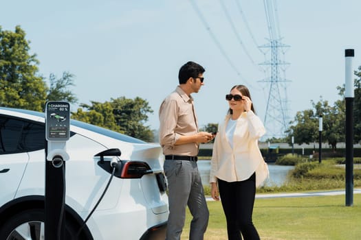 Young couple recharge EV car battery at charging station connected to power grid tower electrical industrial facility as electrical industry for eco friendly vehicle utilization. Expedient