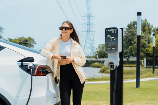 Young woman use smartphone to pay for electricity at public EV car charging station green city park. Modern environmental and sustainable urban lifestyle with EV vehicle. Expedient