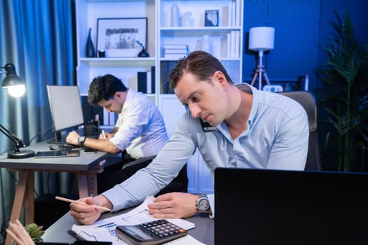 Serious businessman calling on phone with customer to create new project at night time, coworker writing document work sheet on desk. Concept of working on neon blue light modern office. Sellable.