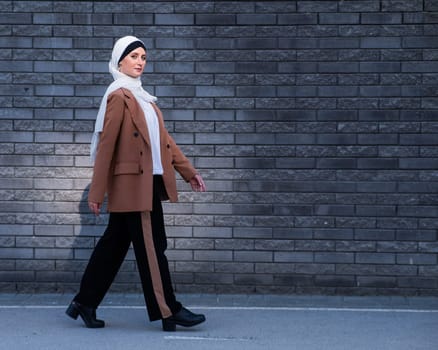 A young woman dressed in a hijab and a business suit walks along a brick wall