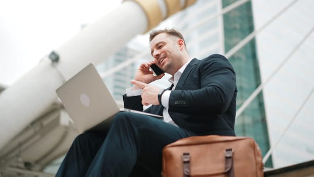 Smart caucasian business man sitting at stairs while using laptop to type to project manager and calling phone to present and plan marketing strategy surrounded by urban city view. Lifestyle. Urbane.