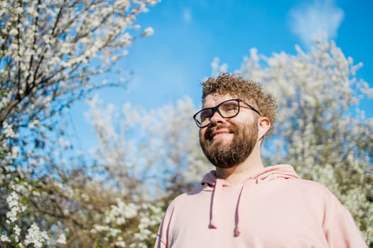 Male bearded guy standing under branches with flowers of blooming almond or cherry tree in spring garden. Spring blossom. Copy space