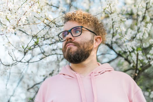 Male bearded guy standing under branches with flowers of blooming almond or cherry tree in spring garden. Spring blossom. Copy space