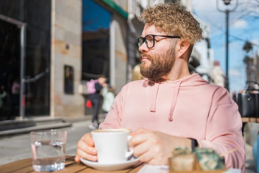 Millennial bearded man having breakfast at table of street cafe on spring day, drinking warm cappuccino. Spring restaurant terrace.