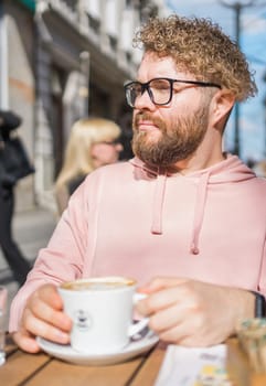 Millennial bearded man having breakfast at table of street cafe on spring day, drinking warm cappuccino. Spring restaurant terrace.