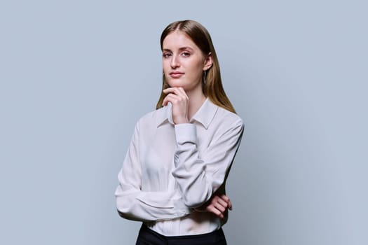 Portrait of young teenage serious female in white shirt with crossed arms on grey studio background. Confident beautiful girl college student looking at camera