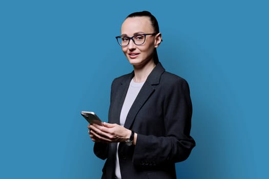 Adult smiling woman holding smartphone on blue background. 30s female looking at camera with phone in hands. Technologies mobile applications internet work business leisure communication