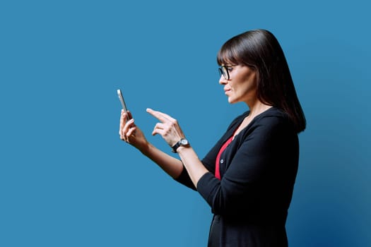 Profile view middle aged serious woman using smartphone on blue background. Mature female looking at phone in hands. Technologies mobile apps applications internet work business leisure communication