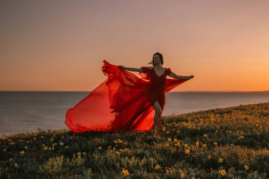 woman red dress is standing on a grassy hill overlooking the ocean. The sky is a beautiful mix of orange and pink hues, creating a serene and romantic atmosphere