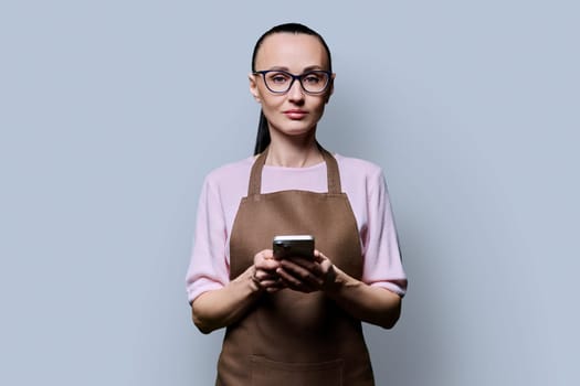 Portrait of 30s woman in apron with smartphone looking at camera on grey background. Serious female using mobile phone texting receiving sending order. Technologies applications service small business