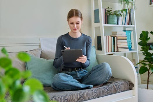Young female teenager with digital tablet, stylus drawing illustrating sitting on couch at home. Technologies, leisure, creativity, education, freelance work, lifestyle, youth concept