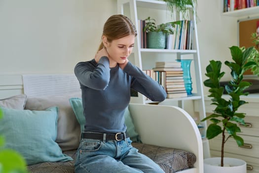 Young teenage female experiencing neck pain sitting on couch at home. Symptoms of the musculoskeletal system, neurological pathologies, acute infectious diseases