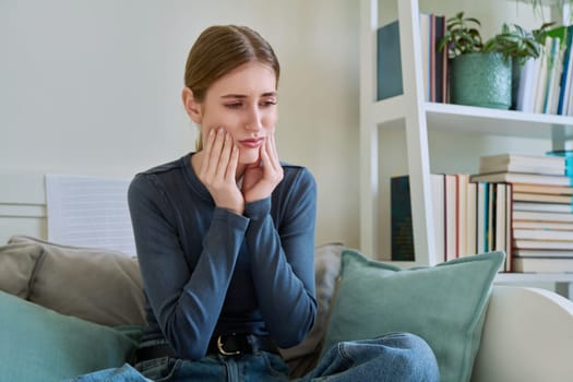Young teenage female having toothache, suffering sad holding hands on face, sitting on couch at home. Teeth dentistry, dental, health, youth concept