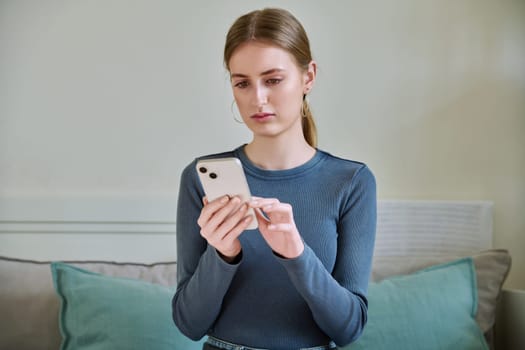 Serious calm female teenager using smartphone, sitting on couch at home, girl 16,17, 18 years old texting reading messages. Modern digital technologies for communication, leisure, learning, shopping