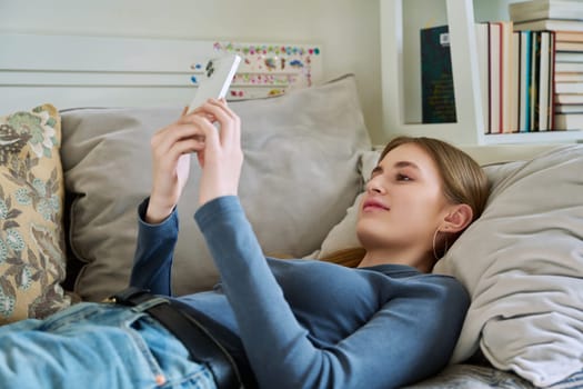 Happy smiling relaxed young female teenager lying on couch at home using smartphone for leisure communication. Youth, lifestyle, digital online technology, gadgets, internet