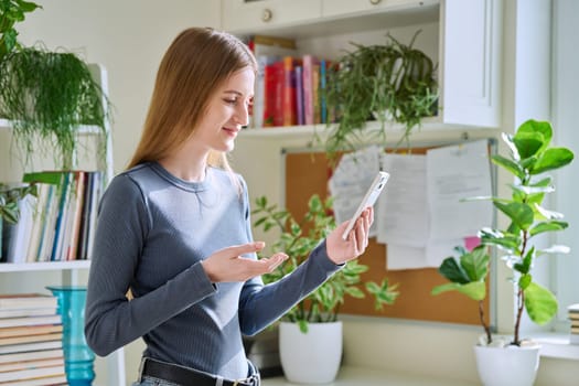 Young teenage female using smartphone for video conversation, home interior. Talking teenage girl looking at the phone screen, online video chat conference call. Technology, communication, leisure