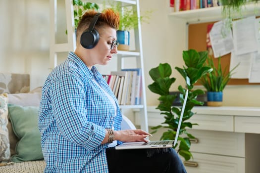 Middle aged woman freelancer in headphones using laptop computer sitting on couch at home. 40s female looking at screen video chat conference call online meeting training mentoring technology for work