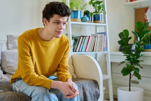 Portrait of handsome smiling guy sitting on couch at home, copy space. Young man in yellow with curly hair, university college student. Lifestyle, youth 19-20 years old concept