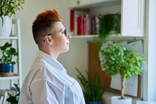 Profile portrait of serious middle-aged woman in glasses with red haircut looking at window in home interior, copy space. Mature people, lifestyle, health, life concept