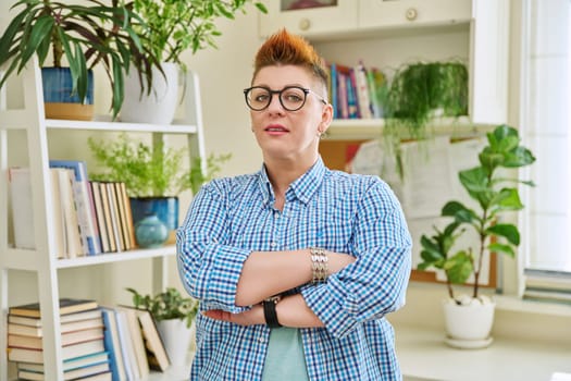 Portrait of smiling confident middle-aged woman in glasses with red haircut, female with crossed arms looking at camera in home interior. Mature people, lifestyle, health, life concept