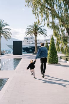 Dad and a little girl are running, holding hands, along the embankment past the fountains. Back view. High quality photo