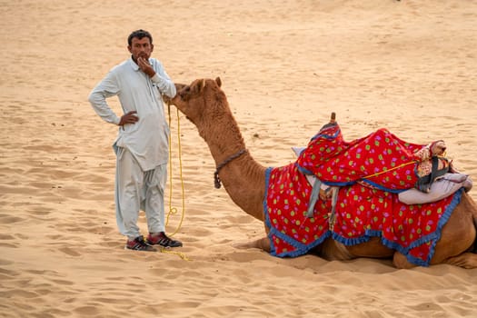 Jaisalmer, Rajasthan, India - 25th Dec 2023: man in traditional kurta pyjama dress standing with sitting camel wearing brightly coloured clothes in the middle of sand dunes