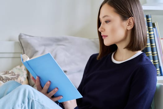 Young woman reading paper book, relaxed sitting on sofa at home. Fiction, hobby, leisure, youth concept