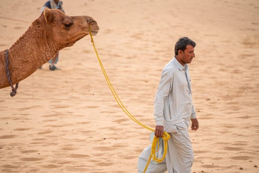 Jaisalmer, Rajasthan, India - 25th Dec 2023:man in traditional kurta pyjama dress leading camel in the middle of sand dunes searching for tourists on a desert safari