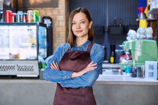 Confident successful young woman service worker in apron with crossed arms looking at camera in restaurant cafeteria coffee pastry shop. Small business, staff, occupation, entrepreneur, owner, work