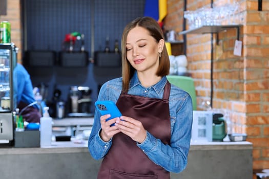 Confident successful young woman service worker in apron holding smartphone in restaurant cafeteria coffee shop pastry shop. Small business, staff, occupation, entrepreneur owner, work concept