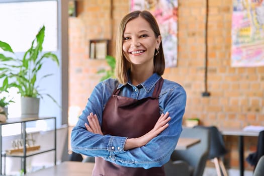 Confident successful young woman service worker owner in apron with crossed arms looking at camera in restaurant cafeteria coffee pastry shop interior. Small business staff occupation entrepreneur work
