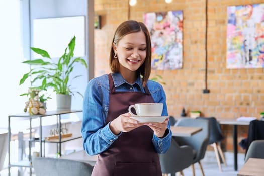 Successful young woman service worker owner in apron with cup of coffee looking at camera in restaurant cafeteria coffee pastry shop interior. Small business staff occupation entrepreneur work