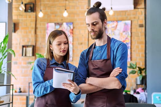 Small business team confident successful colleagues partners owners young man woman in aprons writing at working notebook at workplace in restaurant coffee shop cafeteria. Partnership teamwork work