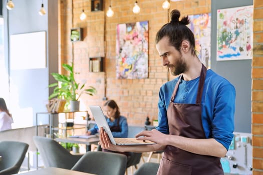 Serious young man service worker owner in apron holding using laptop looking at computer in restaurant cafeteria coffee pastry shop interior. Small business staff occupation entrepreneur work