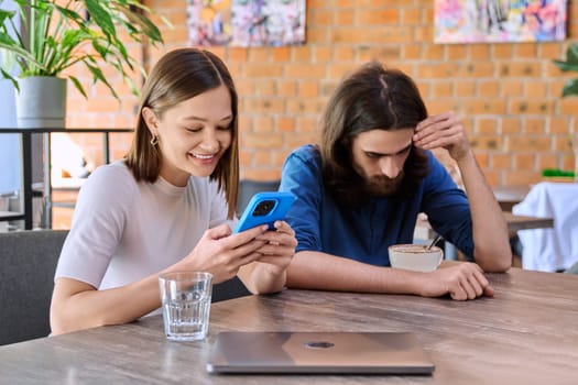 Young handsome people friends, couple man and woman relaxing together in a cafe, using smartphone, drinking coffee. Lifestyle, leisure, youth concept