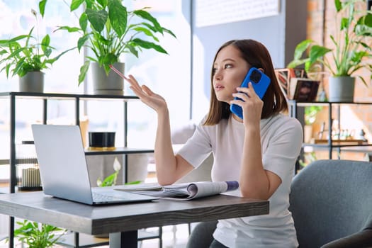 Young woman sitting at table with laptop computer talking on mobile phone in coworking cafe. Female university college student preparing for test exam, freelancer, business employee working remotely