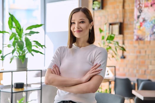 Portrait of young confident beautiful woman with crossed arms looking at camera in coworking cafe. 20s female university college student, businesswoman, freelancer employee, young specialist